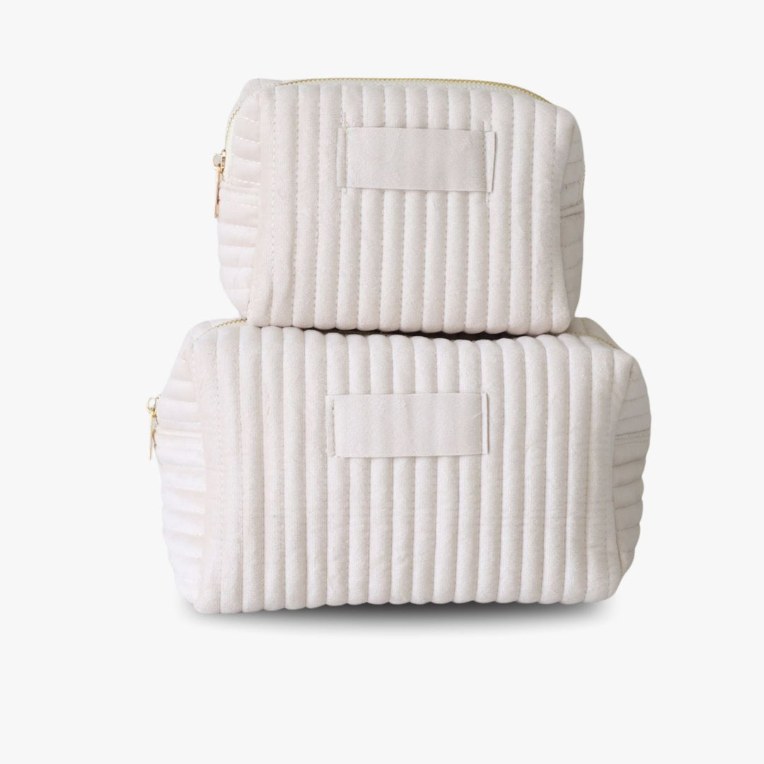 Two nesting cream ribbed velvet toiletry bags with a golden zipper and a tag in the middle for monogramming.