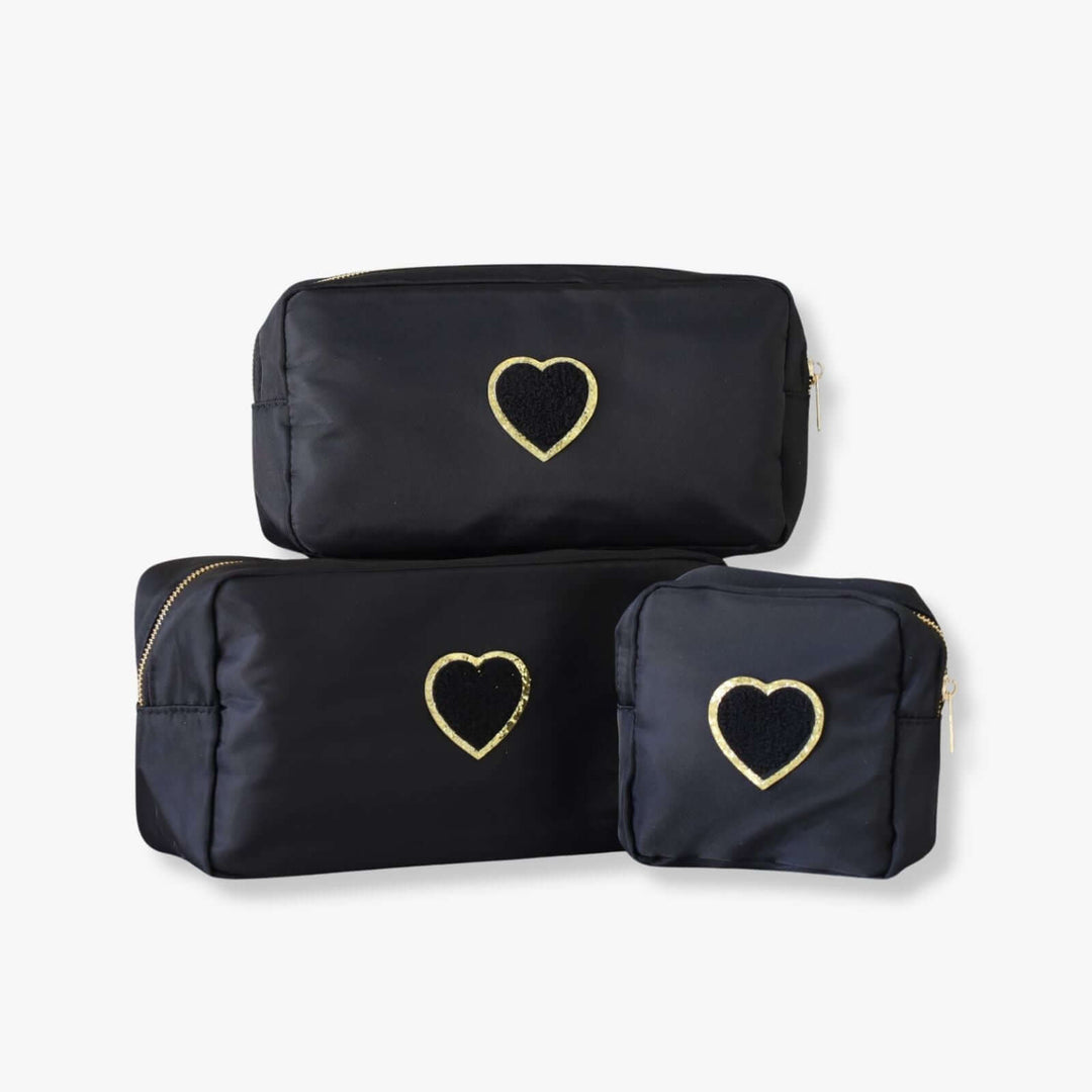 Three black nesting toiletry bags with a heart-shaped patch in the center of each. The patch is black with a gold rim.