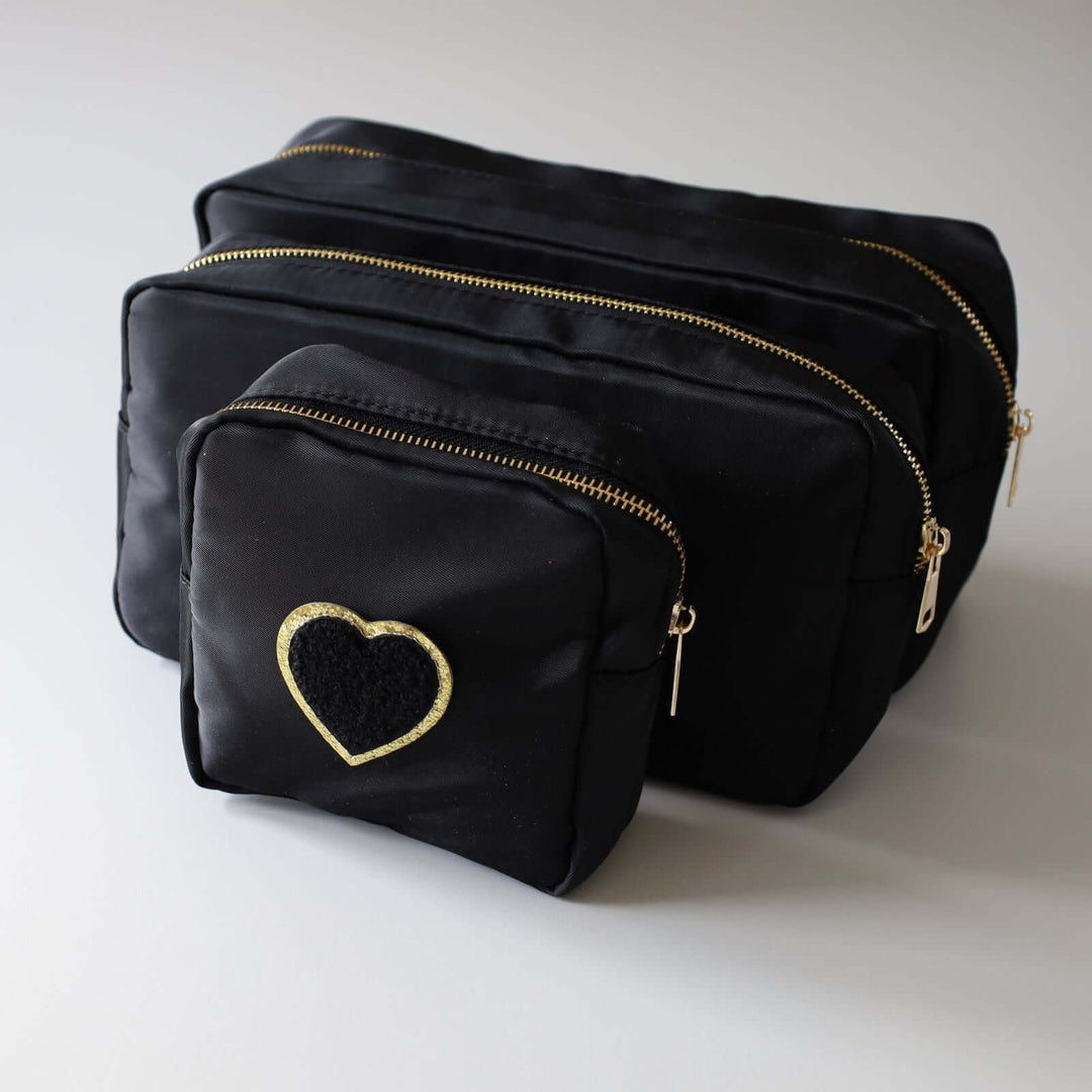 Three black nesting toiletry bags with a heart-shaped patch in the center of each. The patch is black with a gold rim. The zipper's color is gold.