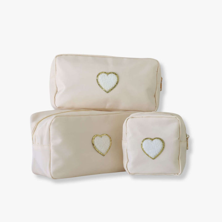 Three ivory nesting toiletry bags with a heart-shaped patch in the center of each. The patch is white with a gold rim.