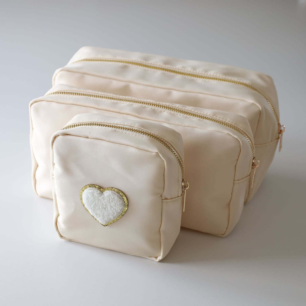 Three ivory nesting toiletry bags with a heart-shaped patch in the center of each. The patch is white with a gold rim. The zipper's color is gold.