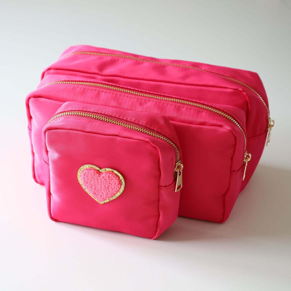 Three pink nesting toiletry bags with a heart-shaped patch in the center of each. The patch is pink with a gold rim. The zipper's color is gold.