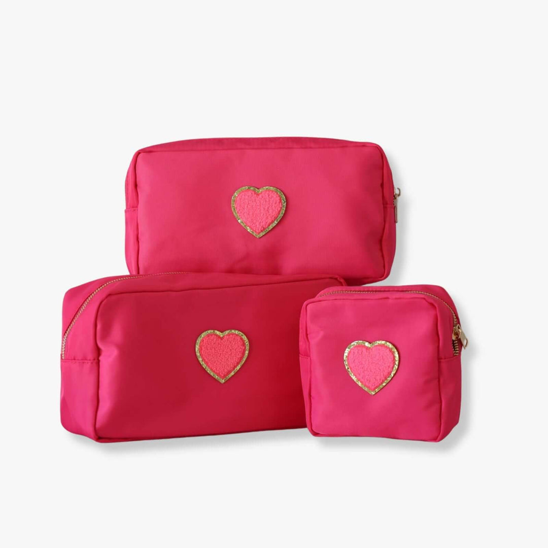 Three pink nesting toiletry bags with a heart-shaped patch in the center of each. The patch is pink with a gold rim.