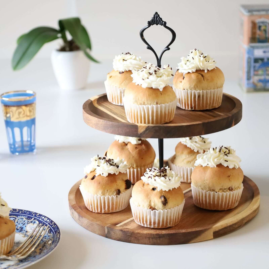 Acacia wood two-tier cake stand with a black metal handle and rod, decorated with buns. There is a white cream on each bun. The cake stand is surrounded with a plate, a box,a small plant,  and a rea glass.