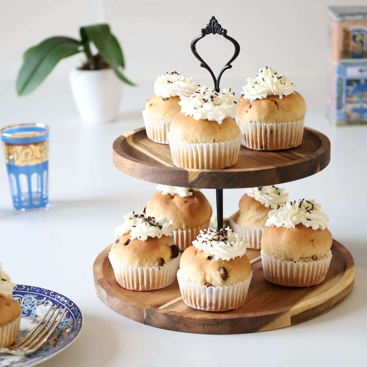 Acacia wood two-tier cake stand with a black metal handle and rod, decorated with buns. There is a white cream on each bun. The cake stand is surrounded with a plate, a box,a small plant,  and a rea glass.