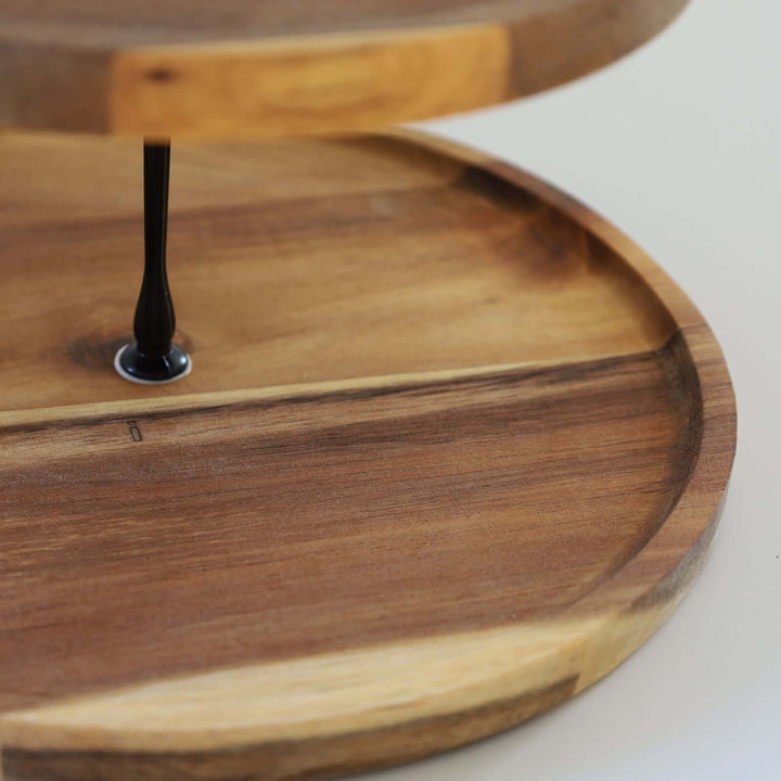 This a a closeup photo of an acacia wood two-tier cake stand with a black metal handle and rod. The wood grain is clearly shown.