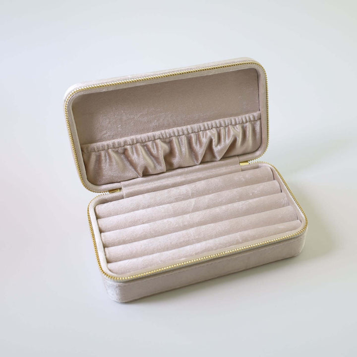 Opened beige velvet jewelry box. Inside we can see multiple slots for earrings, brooches, and rings. There is pocket attached to the lid for necklaces and bracelets