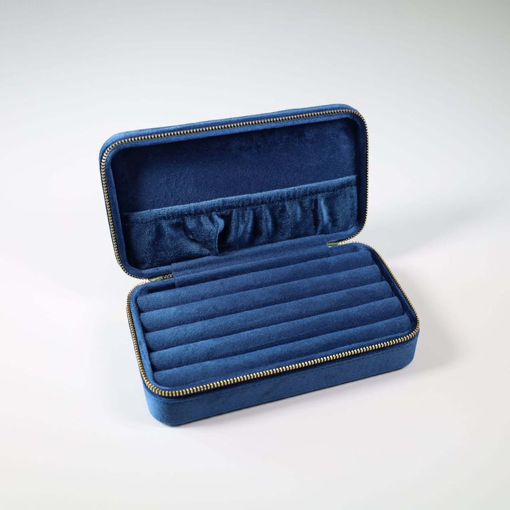 Opened blue velvet jewelry box. Inside we can see multiple slots for earrings, brooches, and rings. There is pocket attached to the lid for necklaces and bracelets.