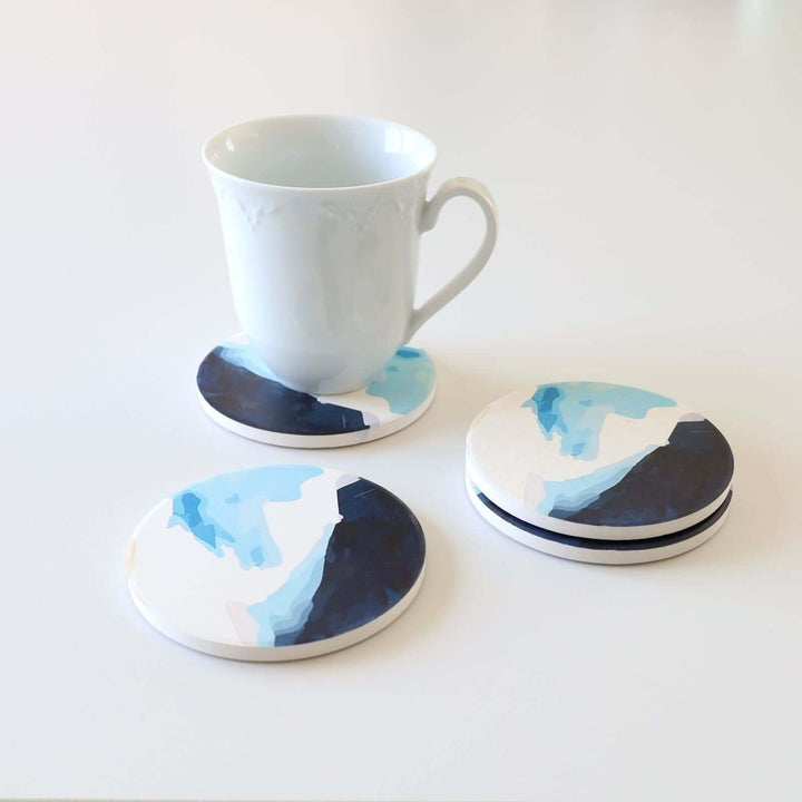 White mug on a round ceramic blue coaster with blue shades that look like waves. There is also the 3 other coasters of the set.