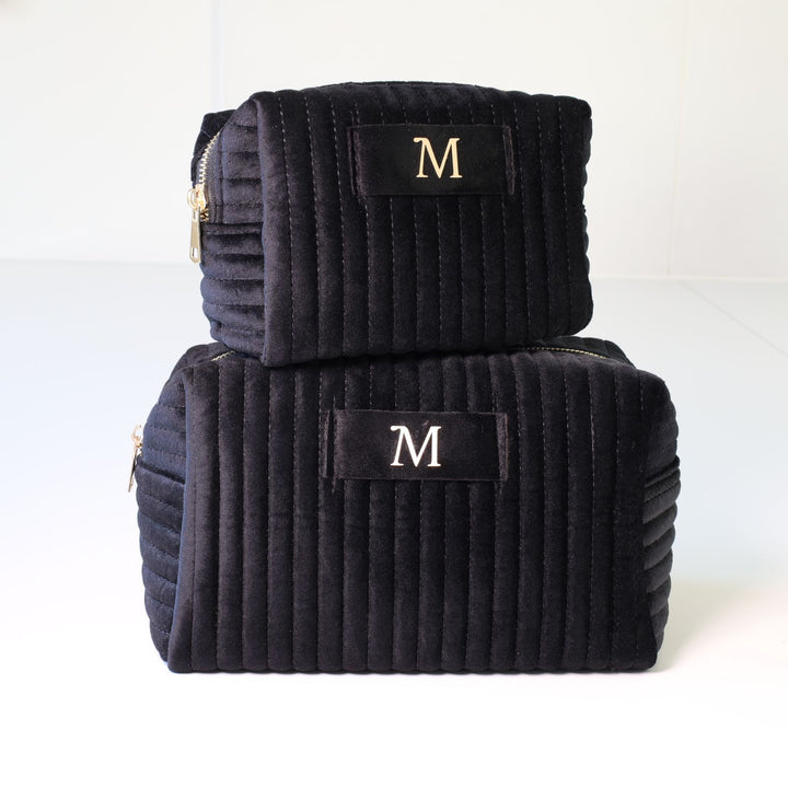 Two nesting black ribbed velvet toiletry bags with a golden zipper and a tag in the middle with a gold foil monogramming.