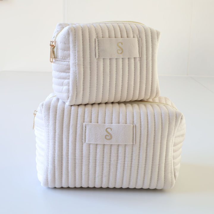 Two nesting cream ribbed velvet toiletry bags with a golden zipper and a tag in the middle with a gold foil monogramming.