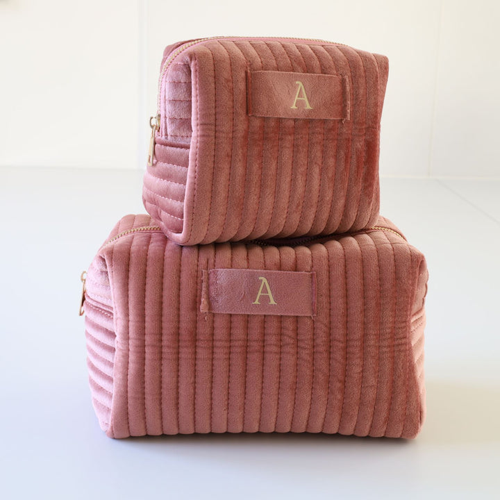 Two nesting pink ribbed velvet toiletry bags with a golden zipper and a tag in the middle with a gold foil monogramming.