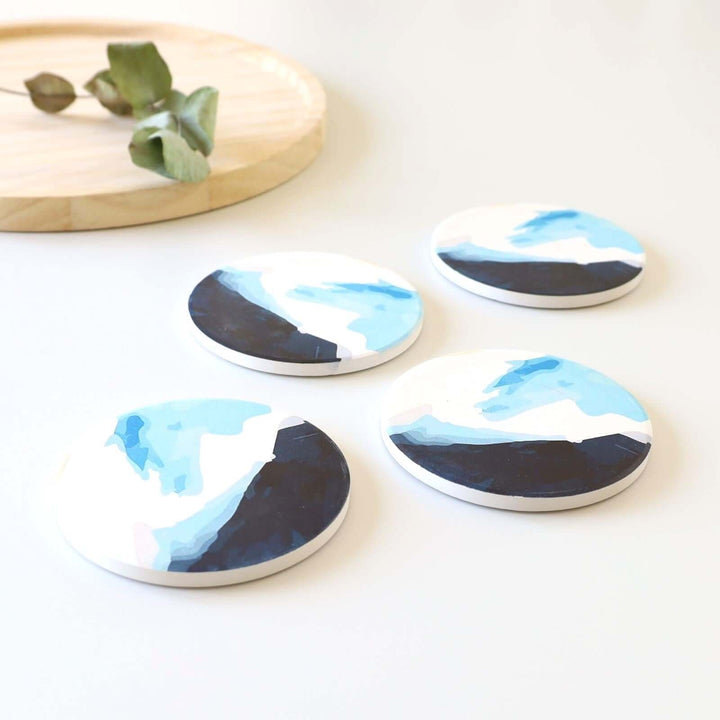 Set of four ceramic blue coasters with a pattern that looks like waves. They are shown on y counter top near a wood tray that holds a dried green plant.