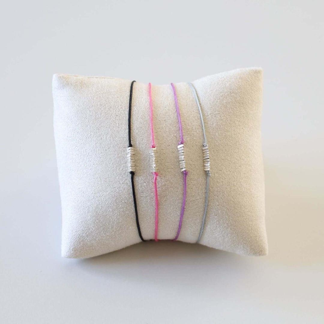 Set of 4 string bracelets in various colors: pink, black, purple and gray. Each bracelet features a string intertwined with small silver bars at the center. The bracelets are shown in a white cushion.