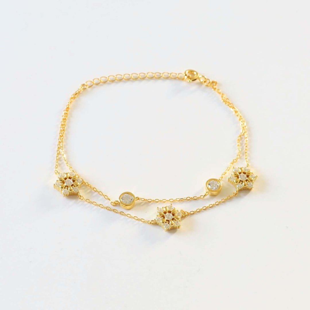Gold plated bracelet featuring a double chain design adorned with three snowflakes, each encrusted with small, sparkling stones, and two round bezel-set stones.