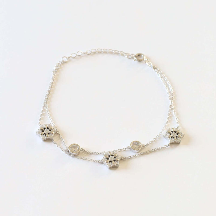 Silver bracelet featuring a double chain design adorned with three snowflakes, each encrusted with small, sparkling stones, and two round bezel-set stones.