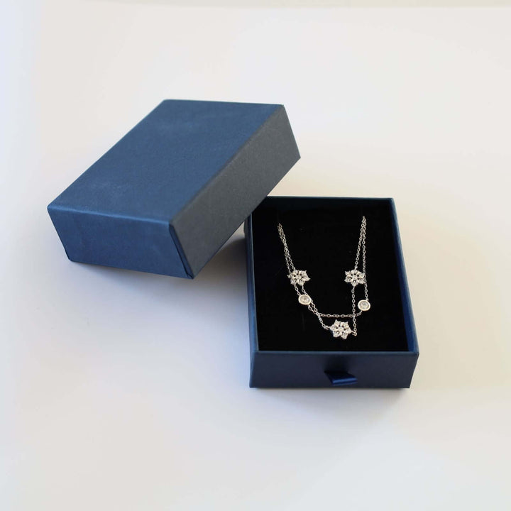 Silver bracelet featuring a double chain design adorned with three snowflakes, each encrusted with small, sparkling stones, and two round bezel-set stones.  The bracelet is shown in a blue cardboard jewelry box.