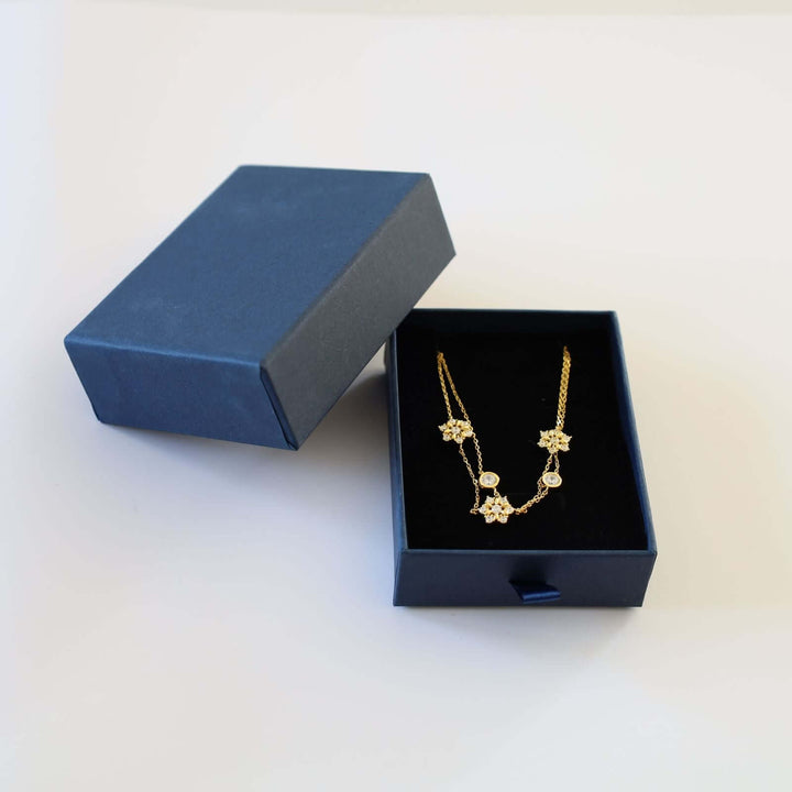 Gold plated bracelet featuring a double chain design adorned with three snowflakes, each encrusted with small, sparkling stones, and two round bezel-set stones.  The bracelet is shown in a blue cardboard jewelry box.