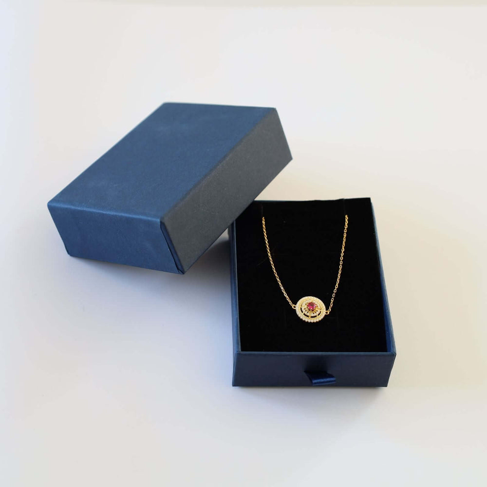 Gold plated bracelet featuring a round charm with a vibrant red stone at the center, encircled by small white stones. The bracelet is shown in a blue cardboard box.