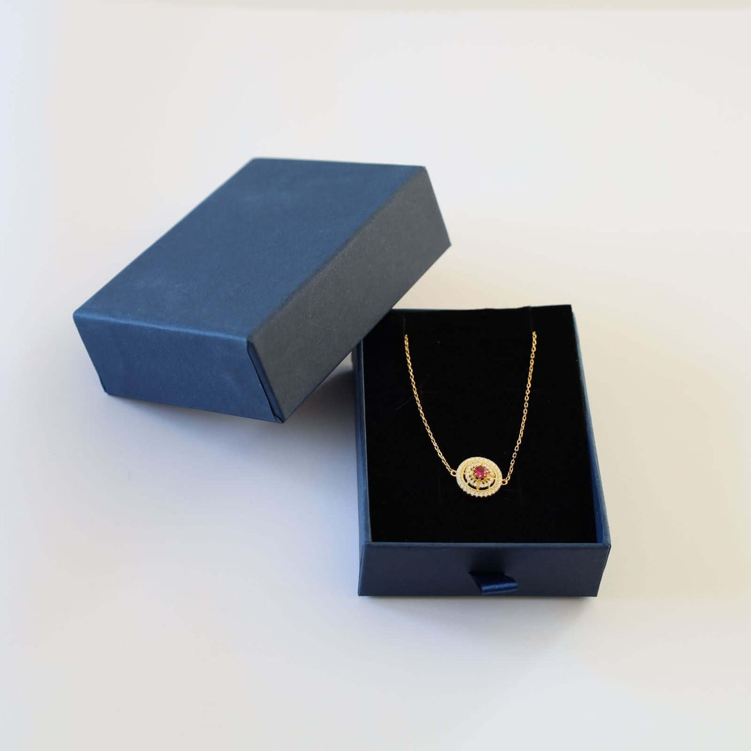 Gold plated bracelet featuring a round charm with a vibrant red stone at the center, encircled by small white stones. The bracelet is shown in a blue cardboard box.