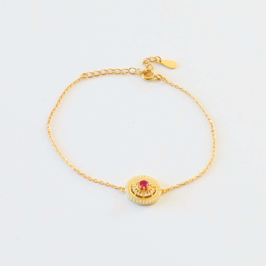 Gold plated bracelet featuring a round charm with a vibrant red stone at the center, encircled by small white stones.