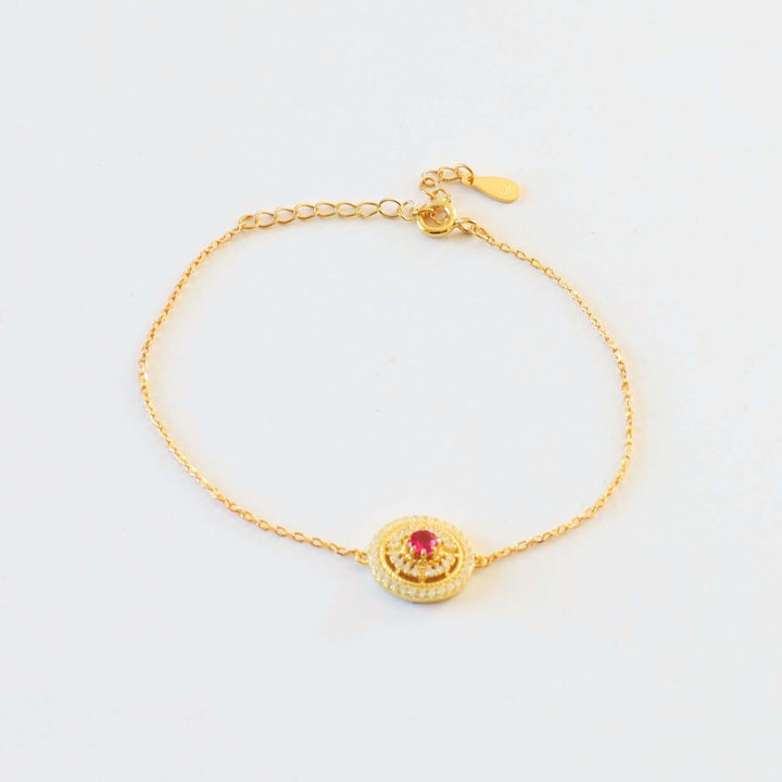 Gold plated bracelet featuring a round charm with a vibrant red stone at the center, encircled by small white stones.