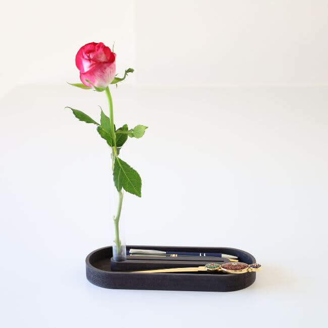 Black pen tray with round edges. The pen tray holds a card holder and a bud vase.