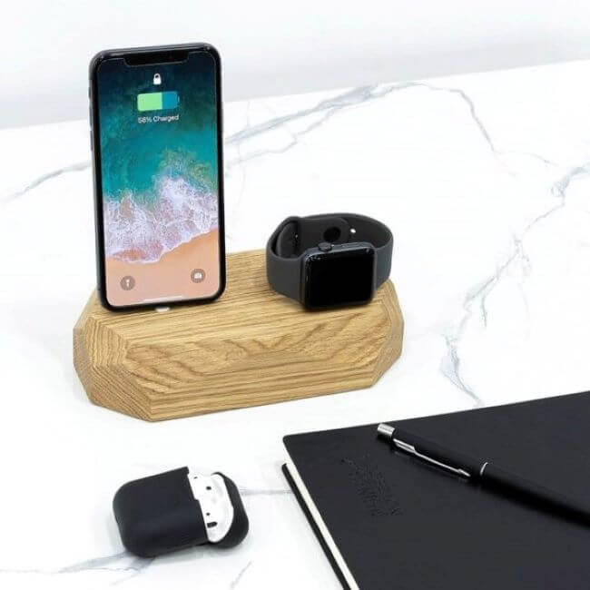 Oak charging dock with an Apple iPhone and a black Apple Watch on a marble desktop next to a black earbud case, a black pen and a black notepad.