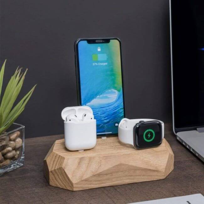 Oak charging station with an iPhone in the middle, a white Apple® Watch  on the right and Airpods in a white case on the left. The  charging station is in the middle of a dark wooden desktop with an open laptop on the right and a plant on the left.