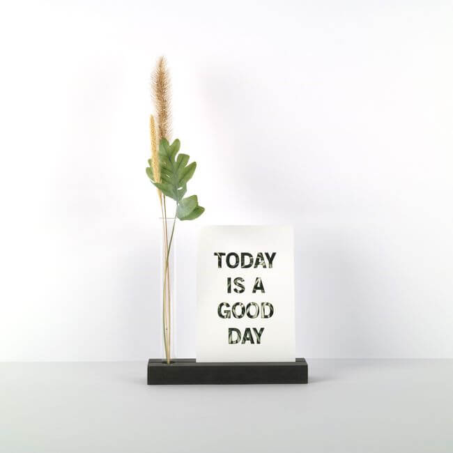 Balck display stand with a card and a bud vase holding a dried flower.