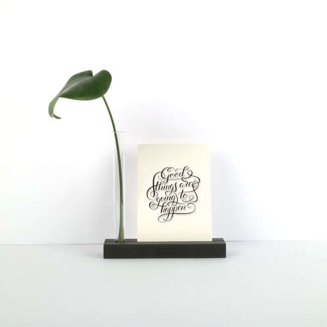 Balck display stand with a card and a bud vase holding a green leaf.