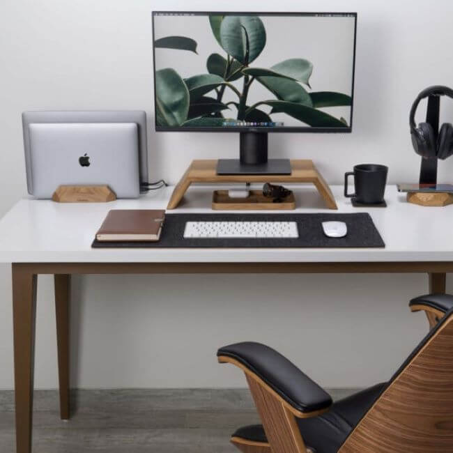 Oak dual vertical laptop stand holding two laptops of different sizes, shown on a desk, in the left-hand side. On the desk there is a headphone stand and a black mug on the right, and a screen on a wood stand in the middle. In front of the screen there is a felt pad holding a brown notepad, a white keyboard and a white mouse. Underneath the screen there is a valet tray holding accessories.