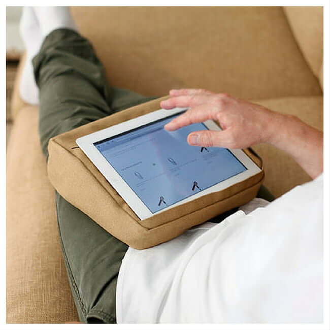 A man surfing on a tablet placed on a beige tablet cushion with pockets for holding and storing the tablet.